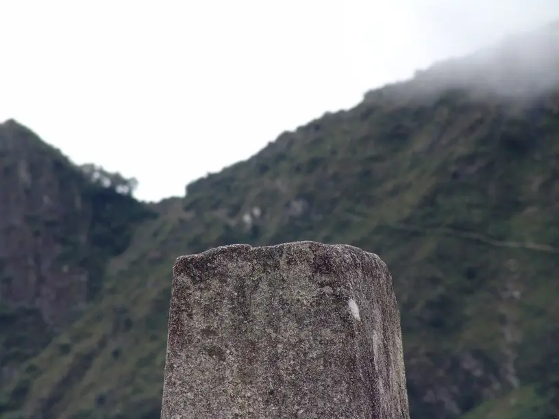 Pilar de pedra cinza e gasta, com o topo plano, diante de um morro verde na névoa.