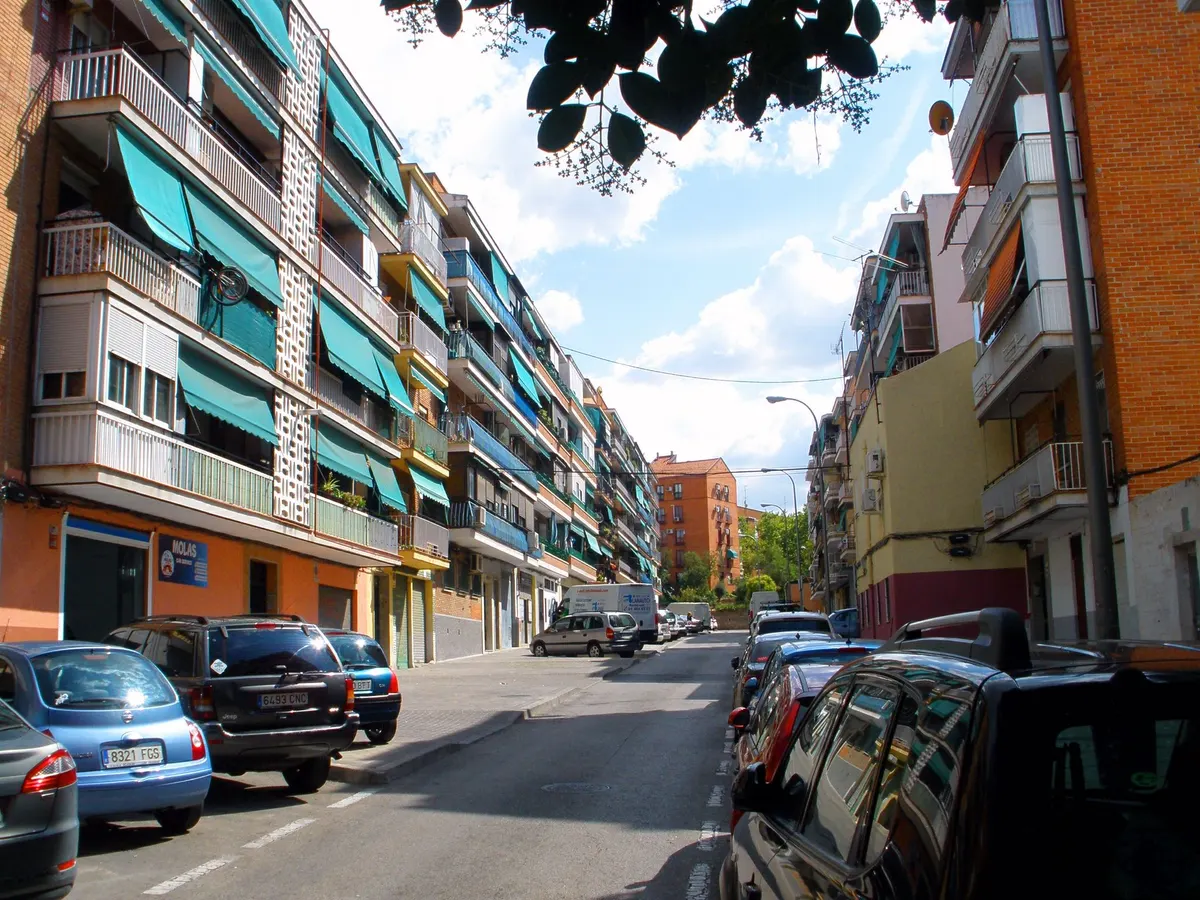 Rua residencial de Entrevías com blocos de apartamentos, varandas, toldos verdes, carros estacionados e galhos no alto.