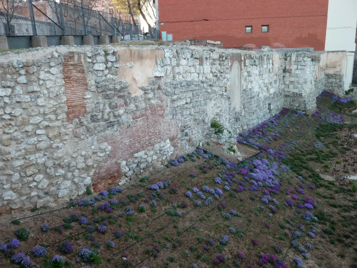 Muralha de pedra com torres retangulares, trechos de tijolo e vegetação junto a um edifício moderno.