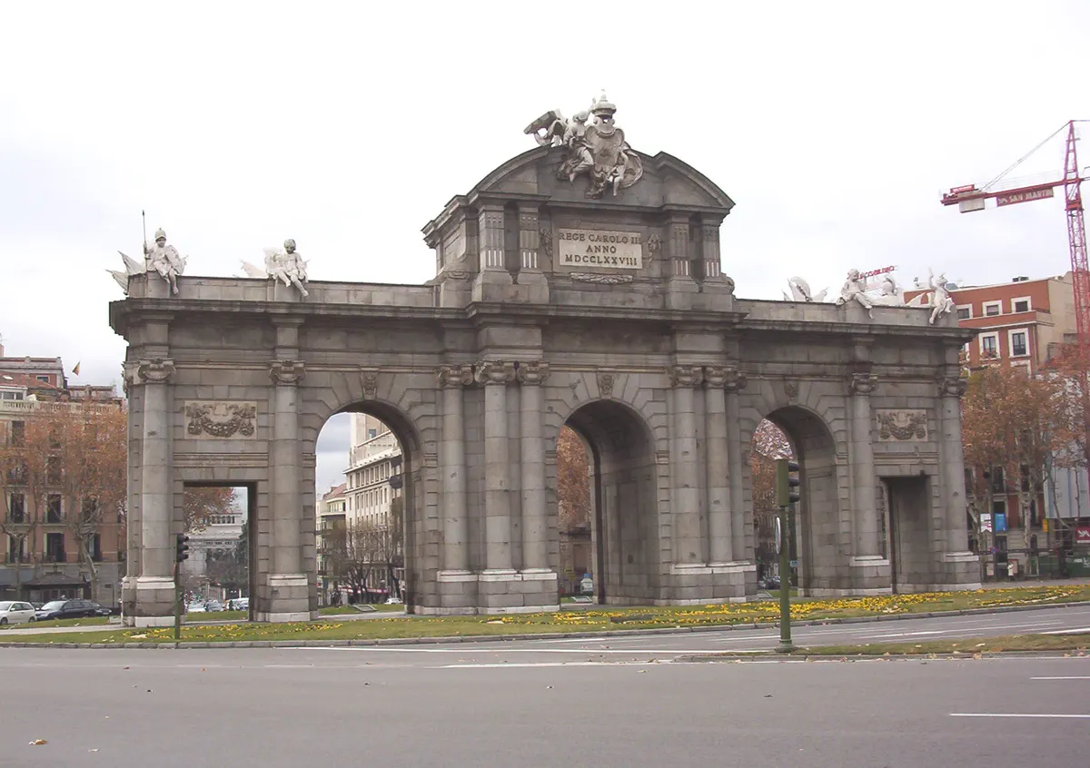 Porta monumental com cinco passagens, colunas, esculturas no alto e inscrição de Carlos III, vista da rua.