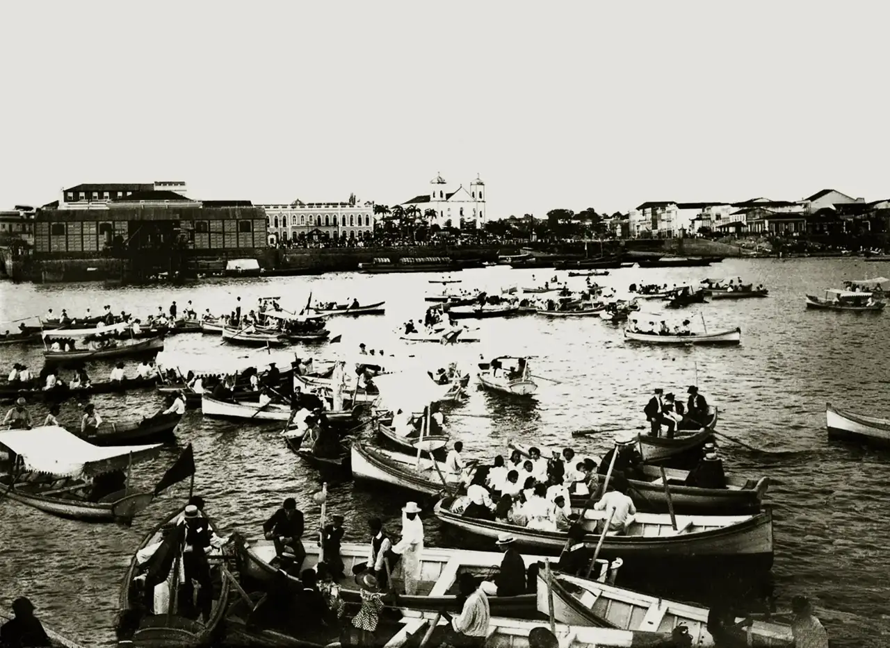 Decenas de barcos y canoas repletos de personas en el río, frente al puerto de Manaos, con una iglesia de dos torres al fondo, en 1902.