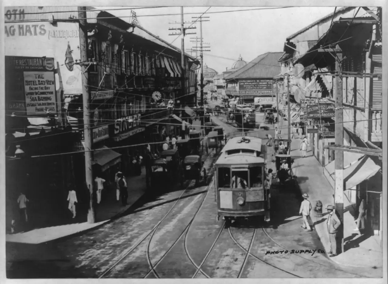Um bonde e automóveis na rua Escolta, ladeada por lojas e cartazes em inglês, com pedestres na calçada, em Manila.