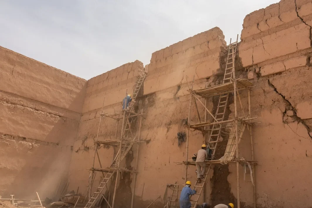 Muralha de terra ocre em reparo, com andaime e pedreiros de costas, sob um céu claro.
