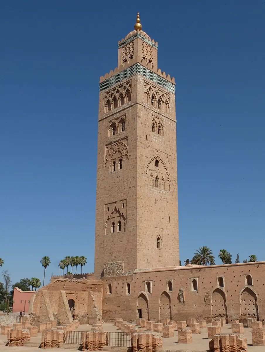Minarete quadrado de pedra com remate dourado, visto da base ao topo, acima de ruínas com pilares baixos, palmeiras e grades.