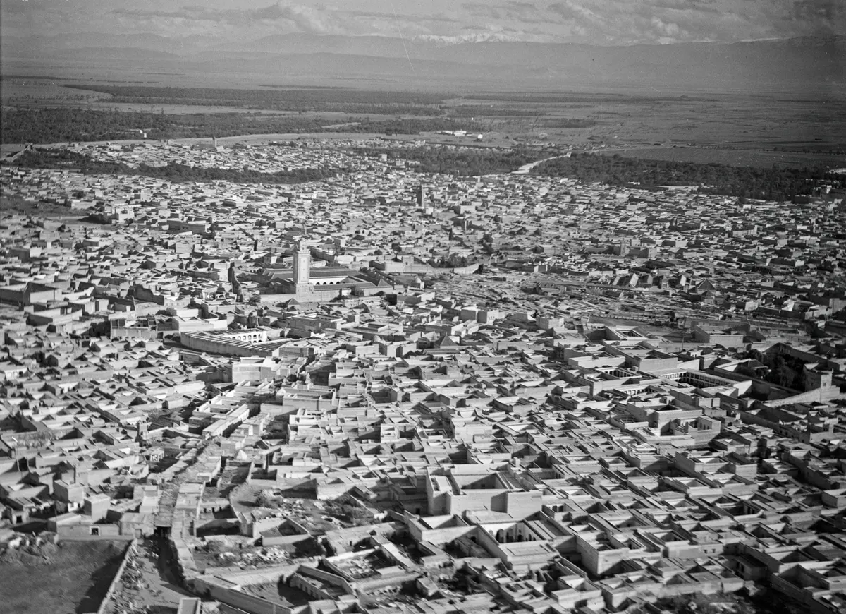 Vista aérea em preto e branco de telhados densos e um minarete, com palmeiras, planície e montanhas ao fundo.