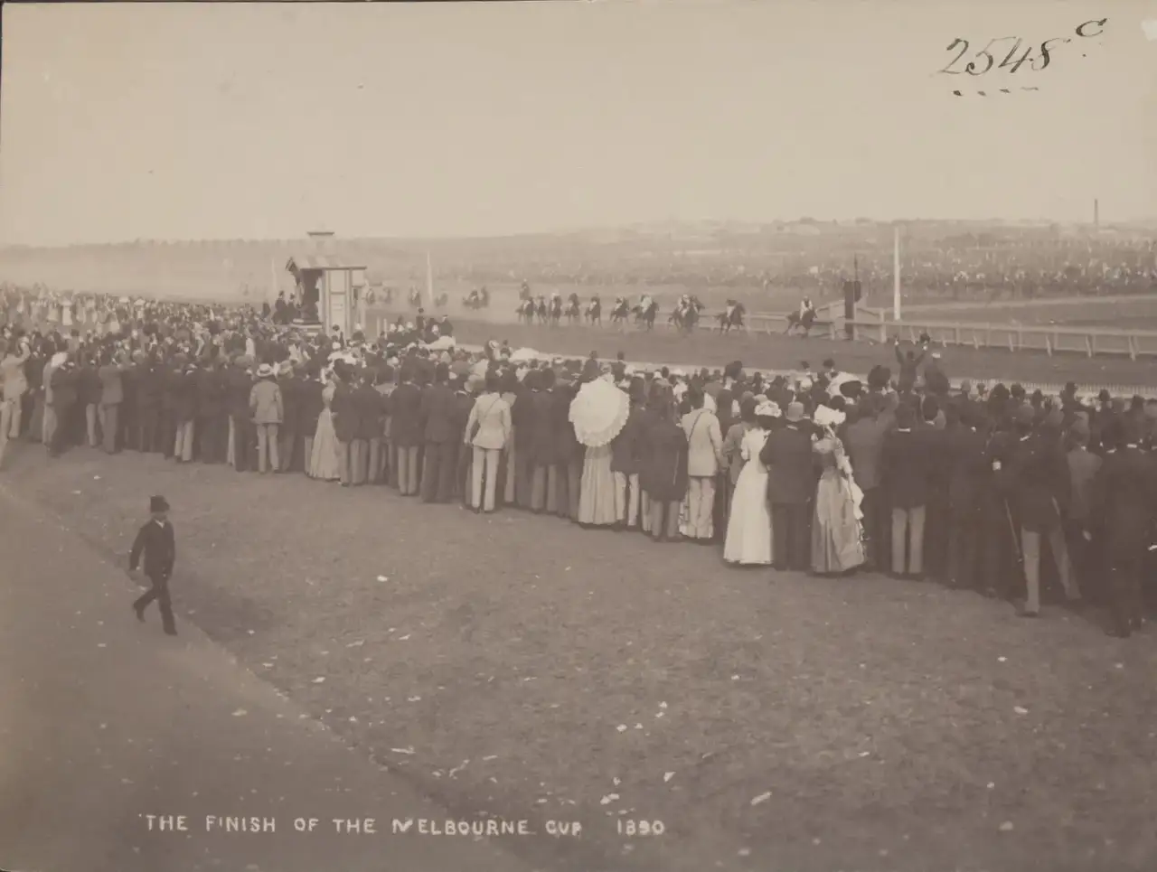 A crowd lined up at the rail watches from behind as horses cross the finish line of the Melbourne Cup race, in 1890.