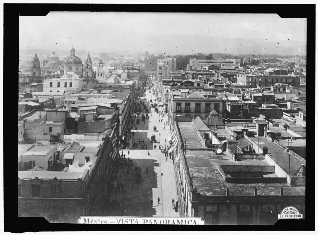 Vista aérea de uma longa avenida reta ladeada por telhados, prédios baixos e torres de igrejas ao fundo, na Cidade do México.