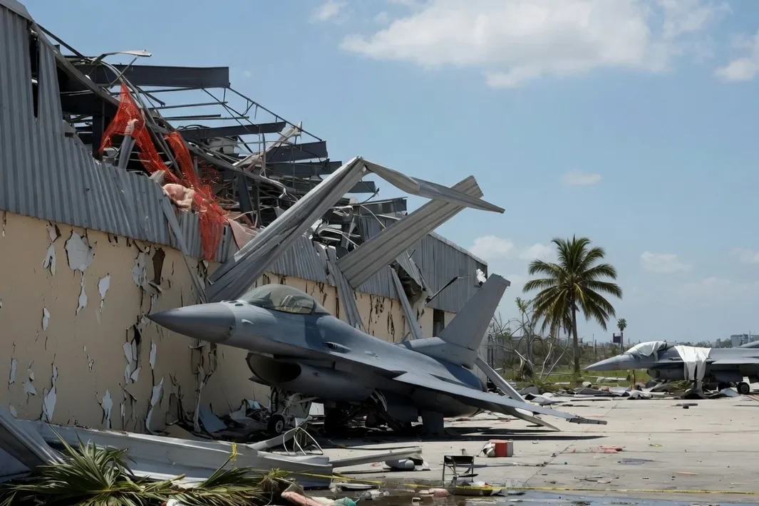 Caza gris con el morro contra la pared de un hangar sin techo, otro avión al fondo y escombros en la plataforma.