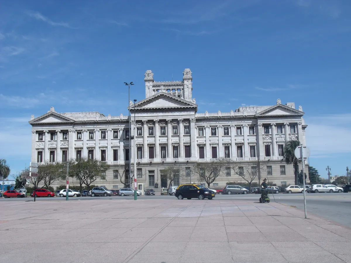 Fachada neoclássica de pedra clara, com colunas, frontão e esculturas no alto, diante de uma praça vazia.