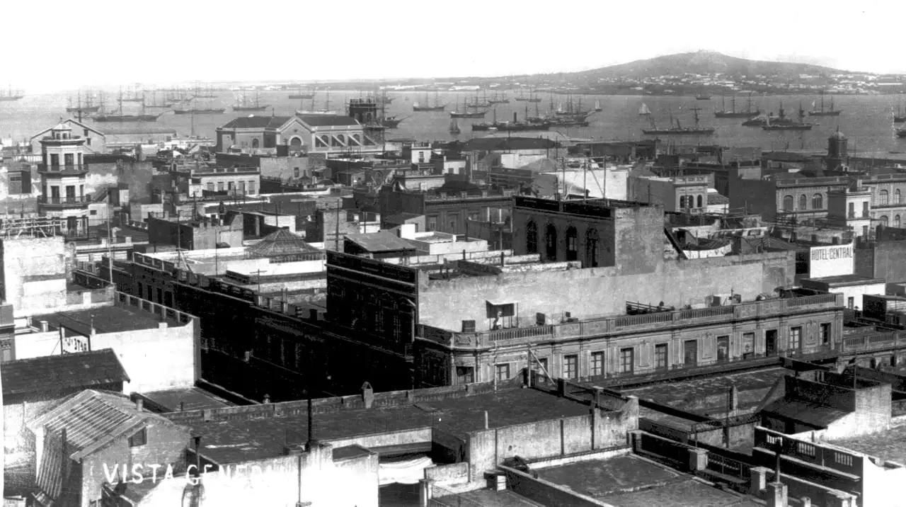 Vista dos telhados do centro de Montevidéu sobre a baía, com o Cerro ao fundo e dezenas de navios de mastros ancorados no porto.