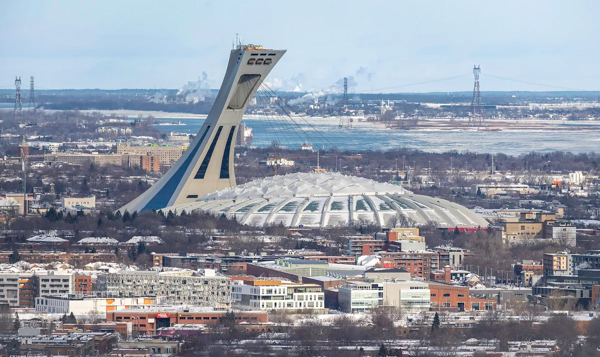 O estádio olímpico de telhado branco e a torre inclinada, vistos de cima, com o rio ao fundo.