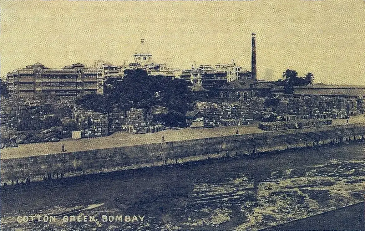 Fardos de algodão empilhados junto ao cais de Cotton Green, com a chaminé de uma fábrica e a cúpula do hotel Taj Mahal Palace ao fundo, em Bombaim.