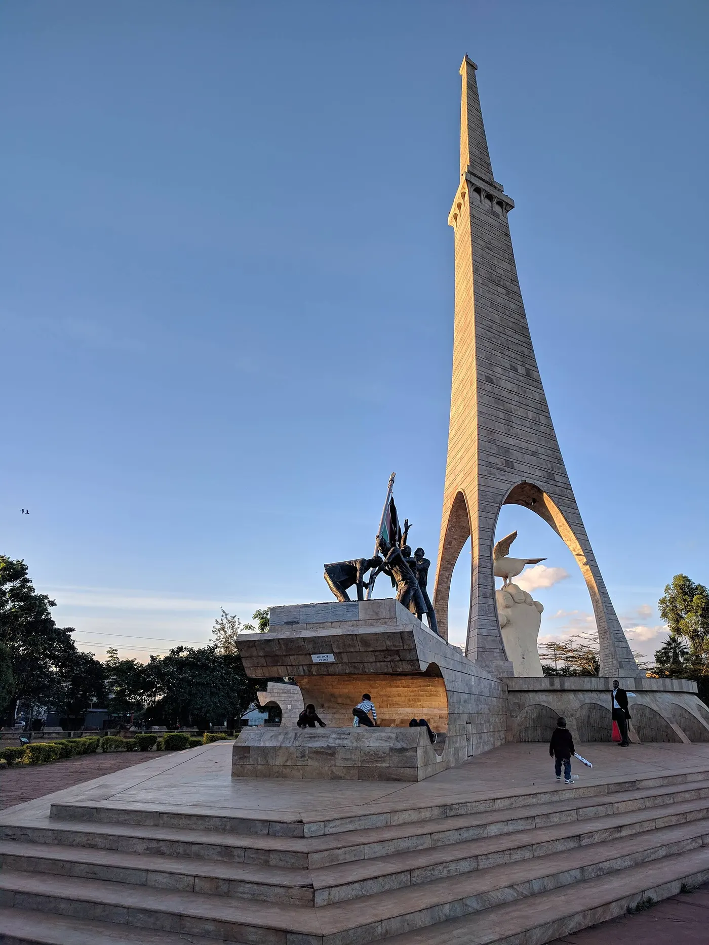 Um obelisco e uma escultura de bronze que ergue uma bandeira, num gramado.