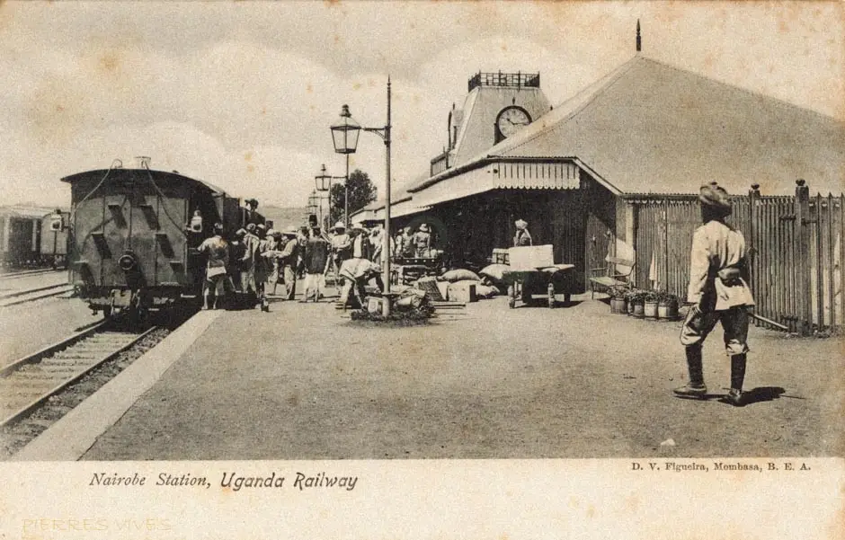 Estação com telhado de duas águas, um vagão na plataforma e pessoas junto aos trilhos.