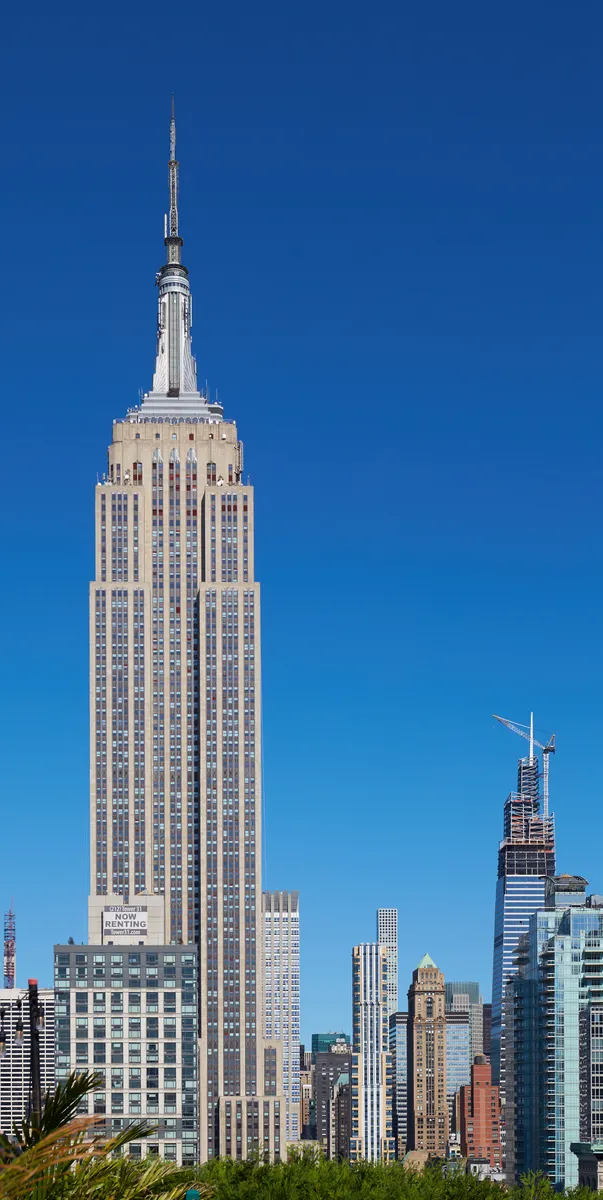 Empire State Building et son antenne sous un ciel bleu, avec immeubles voisins et grue à droite ; des bâtiments au premier plan masquent une partie de la base.
