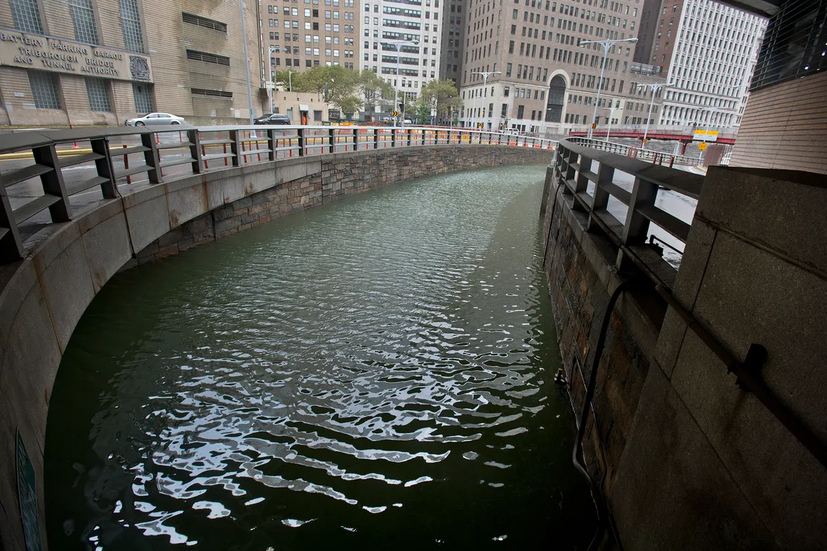 Entrada de túnel inundada, cercada por muros e guardas metálicas, com carros e edifícios ao nível da rua acima.