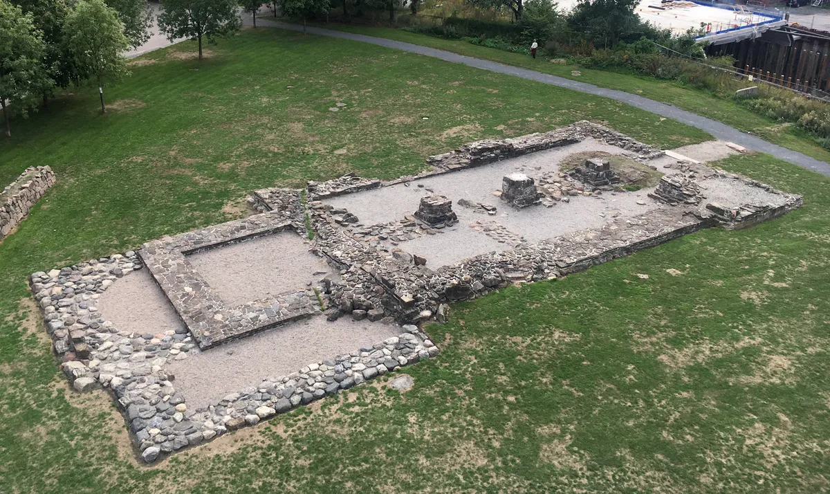 Muros baixos de pedra formando o traçado de uma igreja em terreno gramado, com caminhos, árvores e construções ao fundo.
