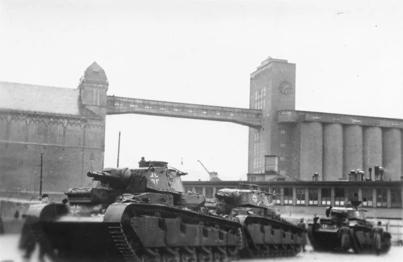 Fotografia em preto e branco de tanques alinhados em rua portuária diante de grande edifício com relógio, com ponte ao fundo.