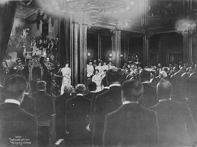 Photographie en noir et blanc d’une cérémonie parlementaire, avec public au premier plan, figures devant un trône et grande peinture au mur.