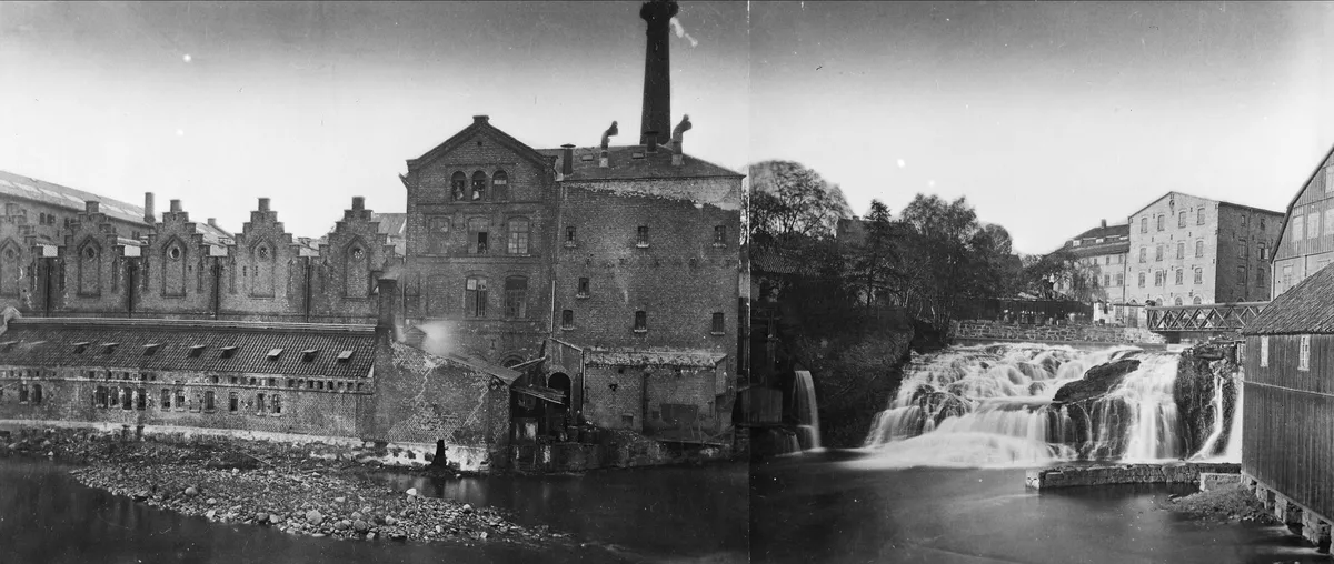 Fotografia em preto e branco de edifícios industriais e chaminés junto a uma queda de água, com passagem sobre o rio e uma junção vertical na imagem.