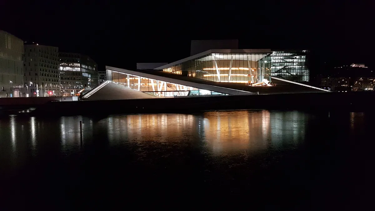 Opéra illuminé de nuit, avec façades vitrées et rampes blanches au bord d’une eau sombre, bâtiments au fond et reflets au premier plan.