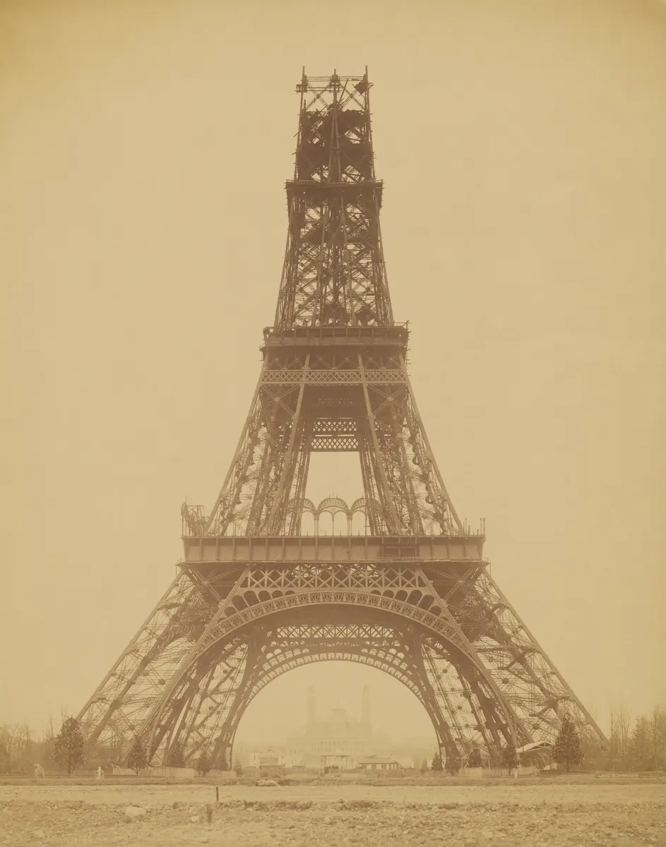 Torre Eiffel incompleta, com dois pisos montados, andaimes e os quatro pilares sobre o terreno da obra.