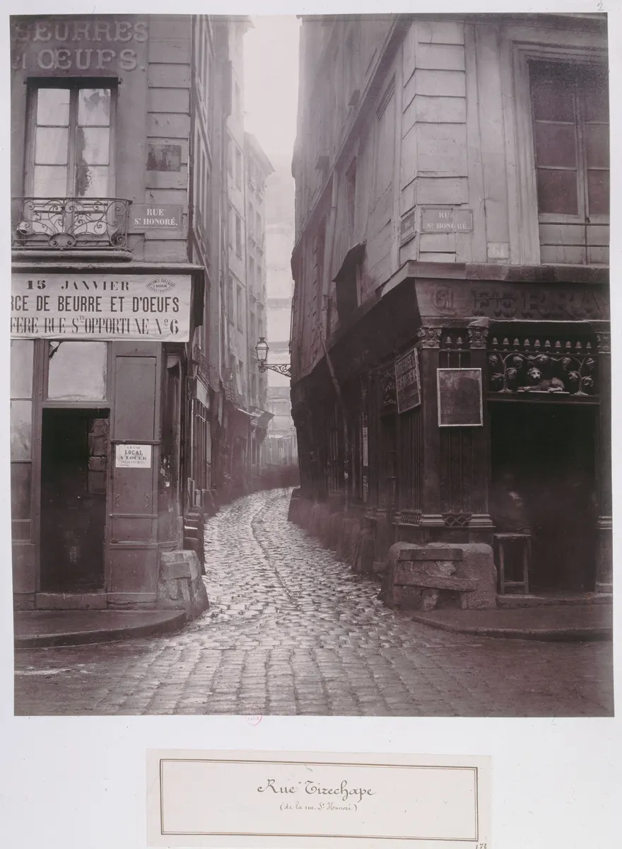 Rua Tirechape estreita e calçada de pedra, entre lojas e edifícios, com etiqueta da montagem original.