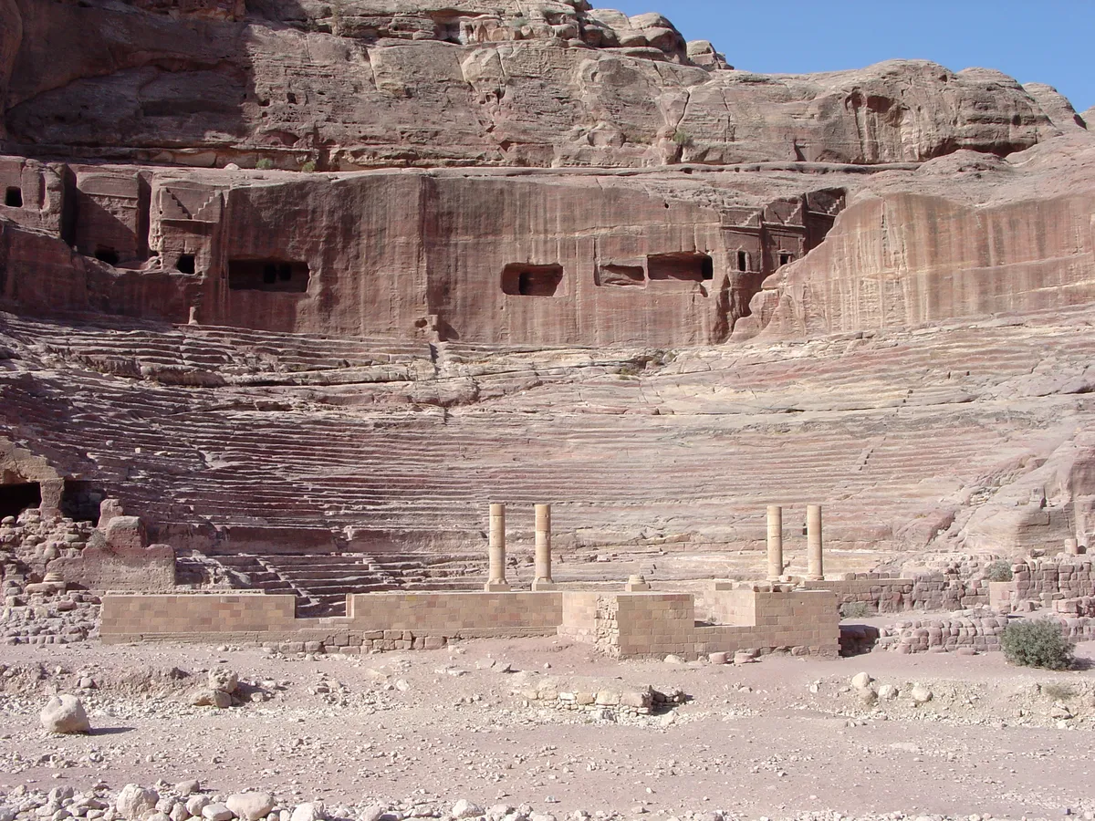 Fileiras semicirculares de assentos talhados na rocha, com aberturas no paredão e ruínas de muros e colunas à frente.