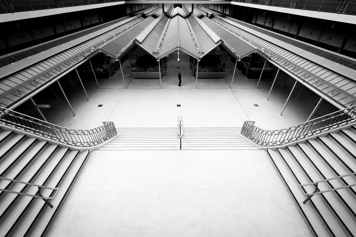 Fotografia em preto e branco do interior do Bolhão, com escadarias simétricas, galerias e coberturas metálicas sobre as bancas, vista do piso superior.