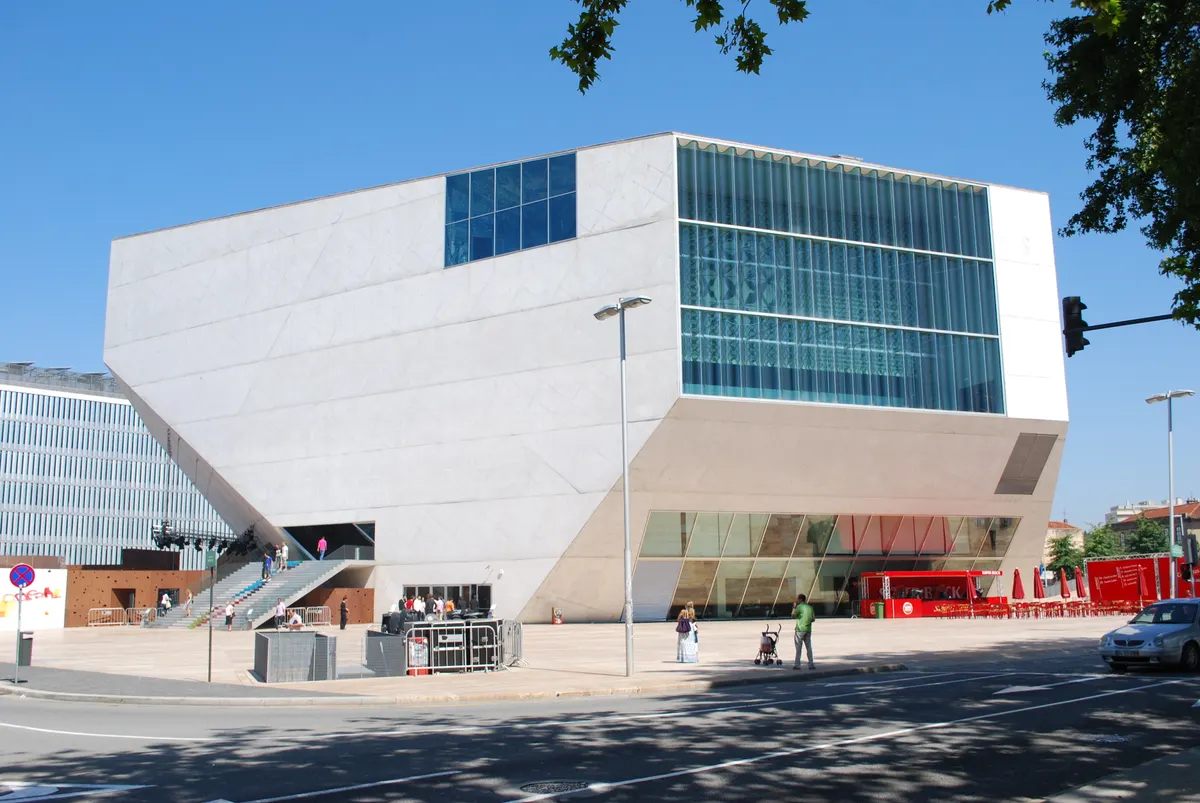 Exterior da Casa da Música sob céu azul, com volume angular claro, fachadas envidraçadas, escadas, praça e rua em primeiro plano.
