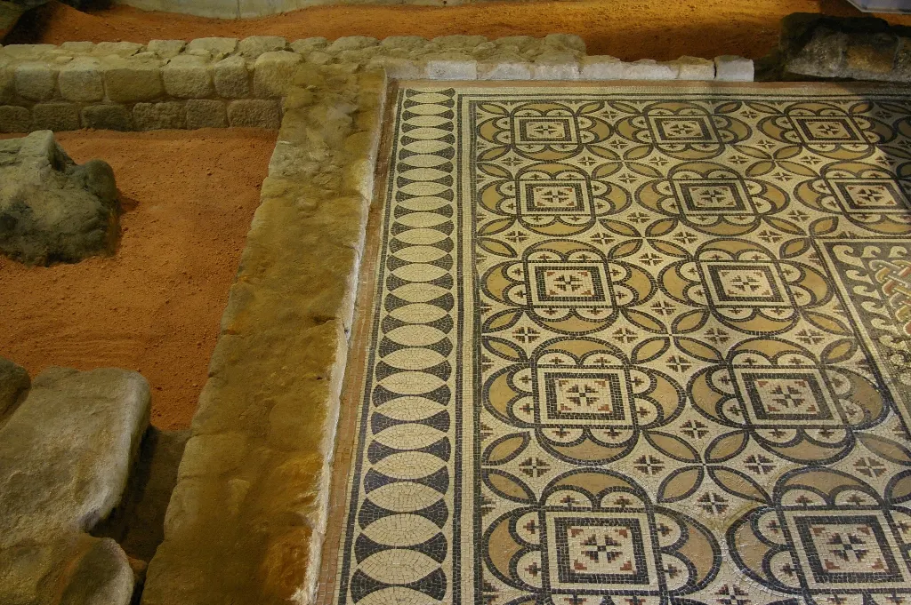 Mosaico de tesselas pretas e brancas com quadrados e laços geométricos, entre estruturas de pedra e terra escavada na Casa do Infante.