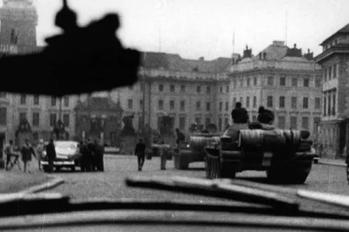 Fotografia em preto e branco feita de dentro de um carro, com blindados e figuras distantes numa praça de Praga; painel e para-brisa em primeiro plano.