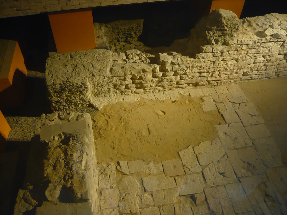 Ruínas arqueológicas de pequenas paredes de pedra sob uma estrutura de cobertura, vistas pela janela pública do Castelo de Praga.