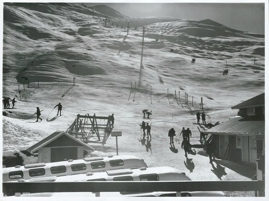 Uma encosta de neve com teleféricos, esquiadores distantes e ônibus brancos na frente.
