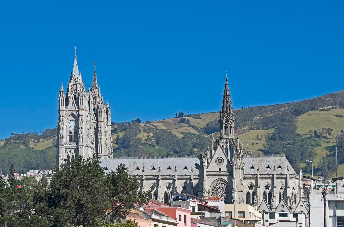 A basílica gótica de pedra cinza, com duas torres e um relógio, diante de morros verdes.
