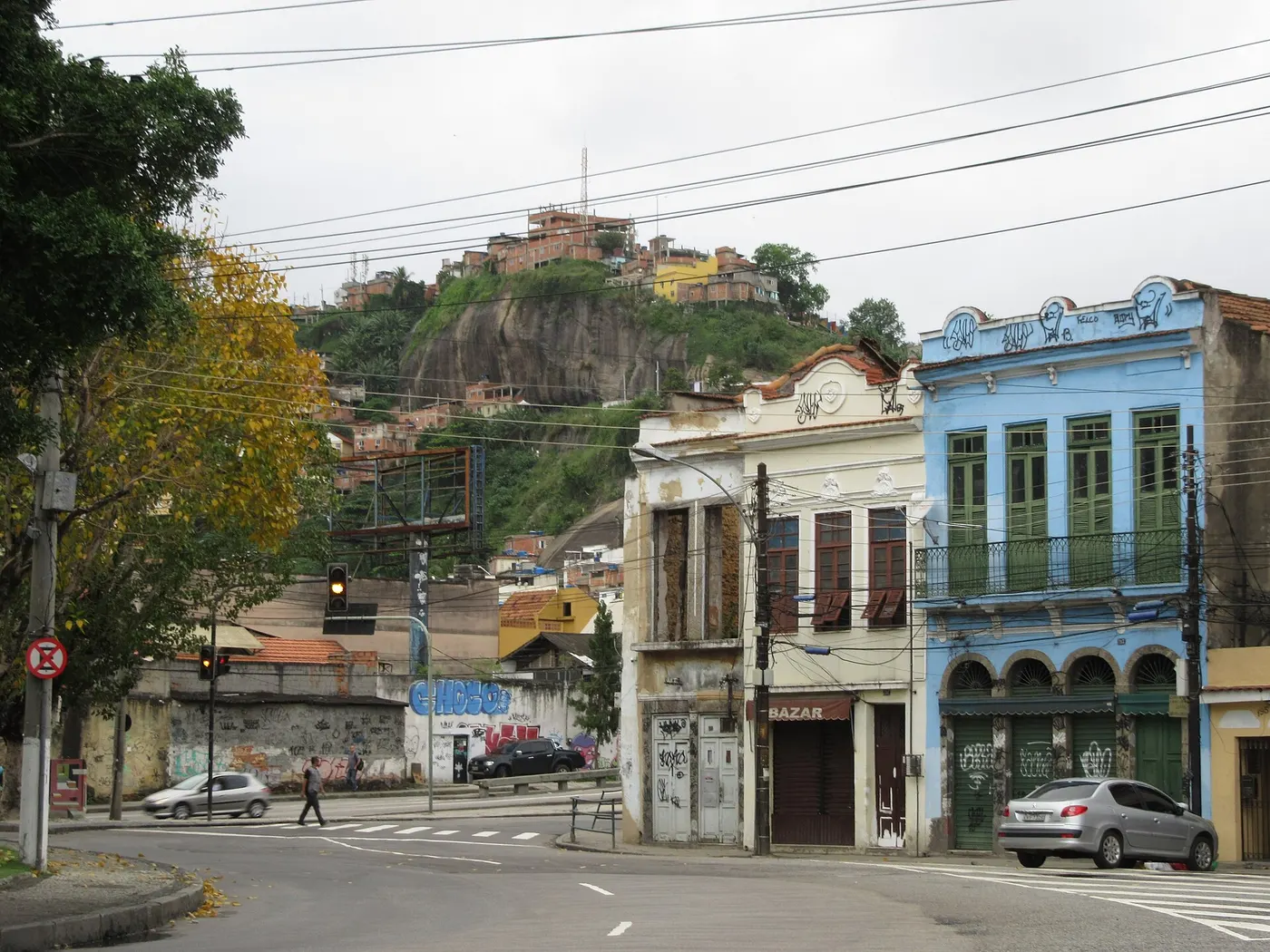 Um morro com casas no alto, visto de uma esquina com sobrados antigos e uma pessoa pequena atravessando a rua.