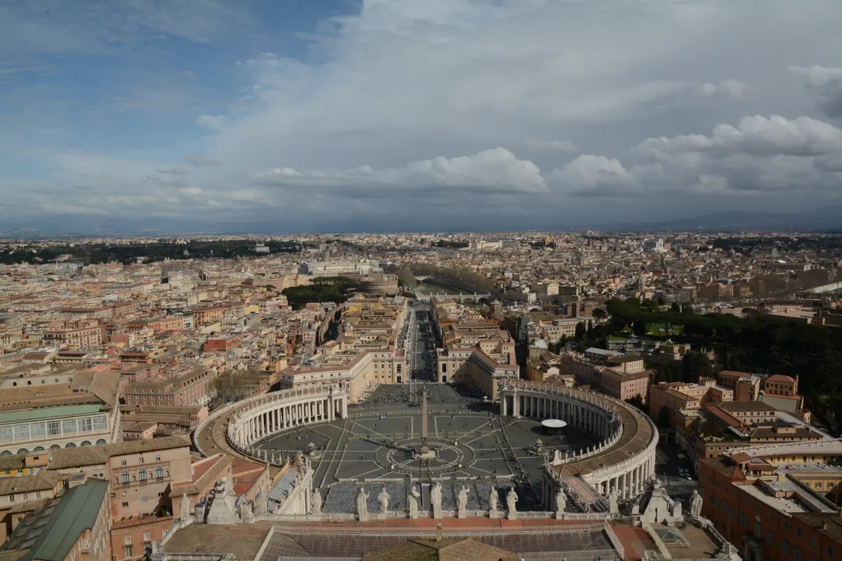 Panorama surélevé de la place Saint-Pierre, colonnades, obélisque, Via della Conciliazione et toits de Rome sous un ciel nuageux.