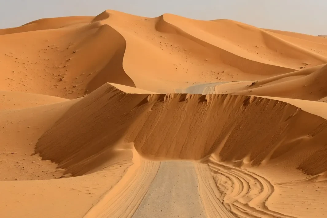 Un talud de arena cruza dunas anaranjadas y corta una huella de vehículo en el desierto.
