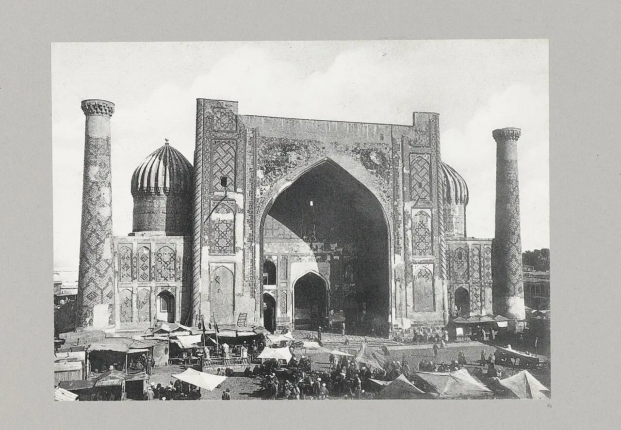 Portal de madrassa com dois minaretes e barracas de mercado na frente.