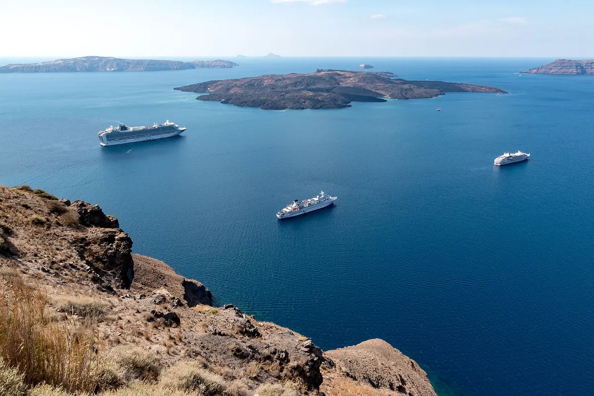 Três navios brancos na água azul, a ilha escura ao fundo e o penhasco em primeiro plano.