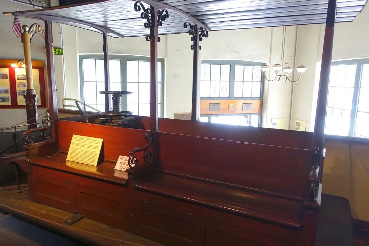 Interior de carro antigo com bancos de madeira, mecanismo de controle, teto, janelas e pequenas placas ao fundo no museu.