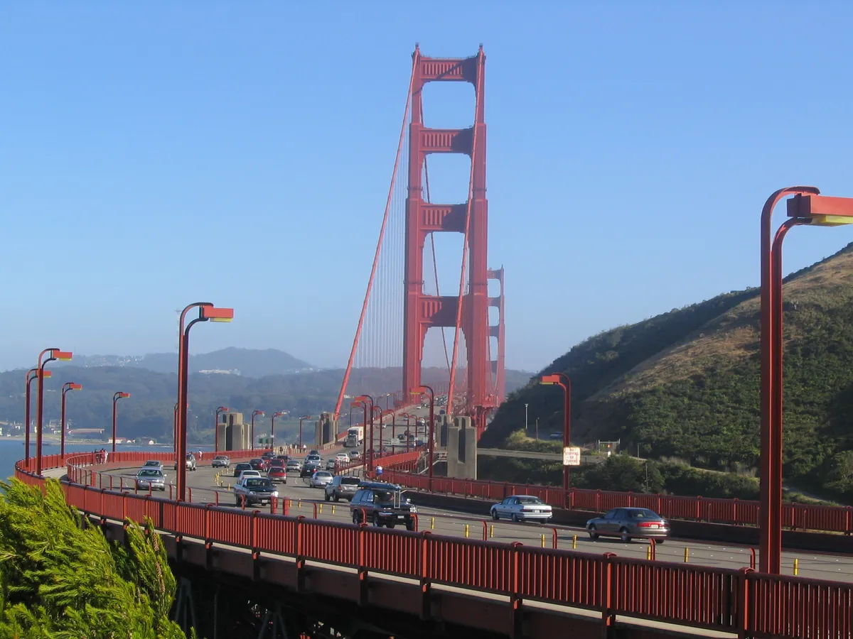 Vista da ponte Golden Gate com torres e cabos vermelhos, pista com carros, postes, passarela e morros ao fundo.