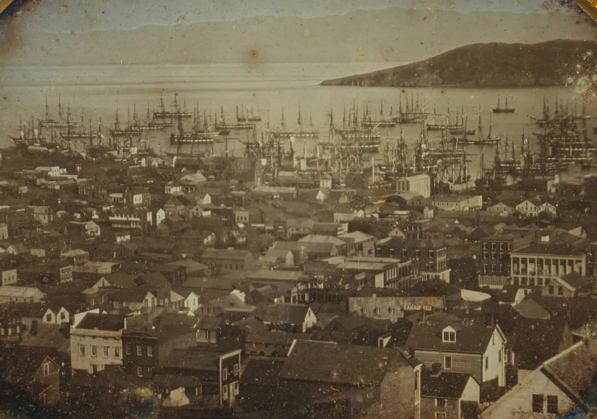 Daguerreótipo em tons dourados com telhados no primeiro plano, muitos mastros e barcos no porto e uma ilha ao fundo, com bordas manchadas preservadas.
