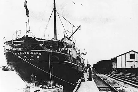 Um navio a vapor escuro atracado junto a um galpão e a trilhos, em fotografia antiga.