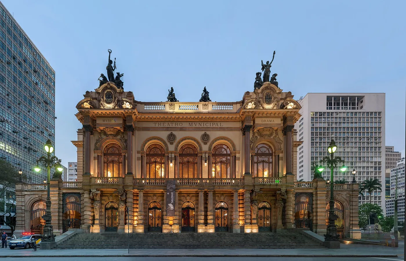 A fachada iluminada de um teatro clássico, com escadaria e torres modernas dos dois lados.