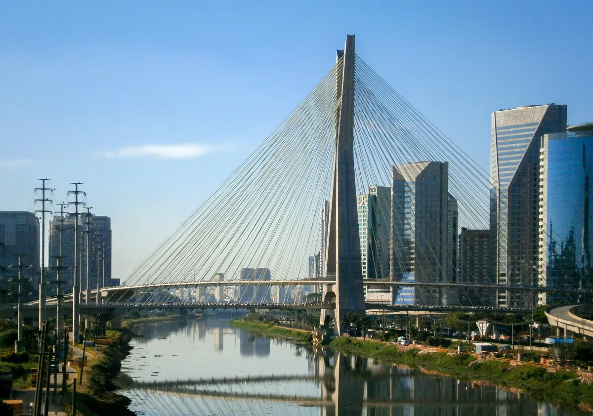 Mastro e cabos da ponte sobre o rio, com torres de vidro ao fundo.