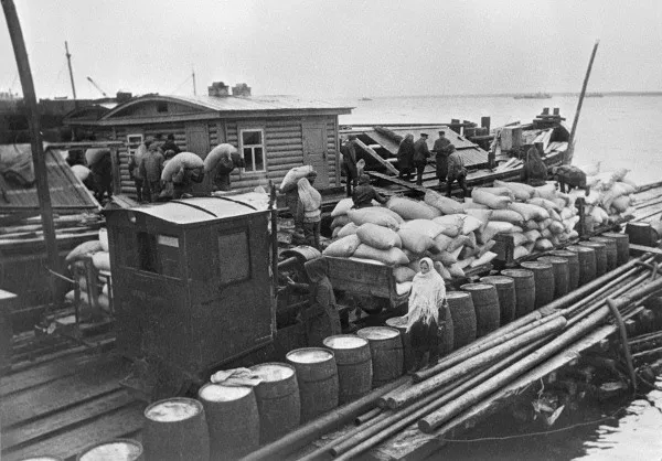 Fotografia de trabalhadores e sacos sobre uma embarcação de carga, com barris, mastro, cordas e água ao fundo.