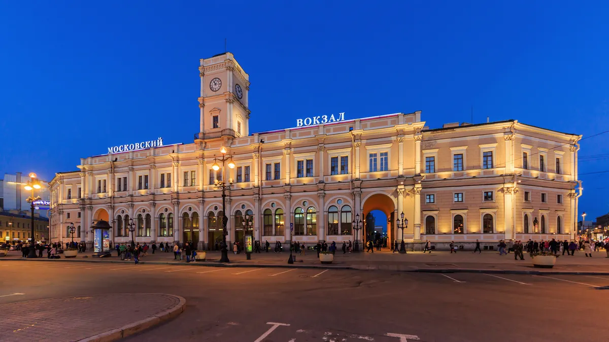 Fachada iluminada da estação com torre e relógio, edifícios laterais, praça, postes e algumas pessoas ao anoitecer.