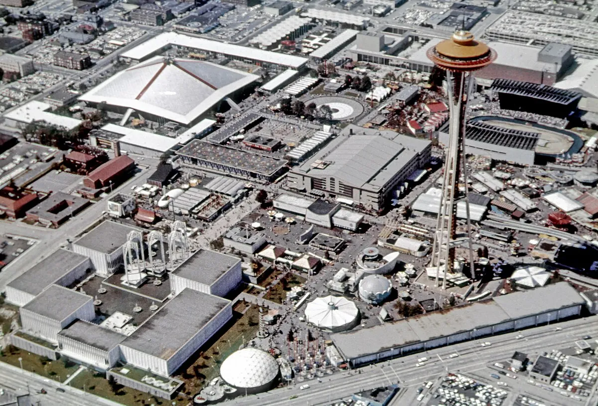 Vue aérienne de l’exposition, avec la tour au disque orange à droite, des pavillons blancs, des arcs et une place circulaire.