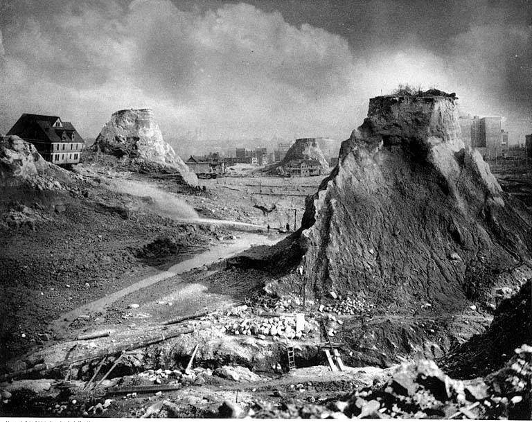 Une colline coupée, avec des maisons sur ce qui reste des crêtes et un jet d’eau au fond de la fouille.